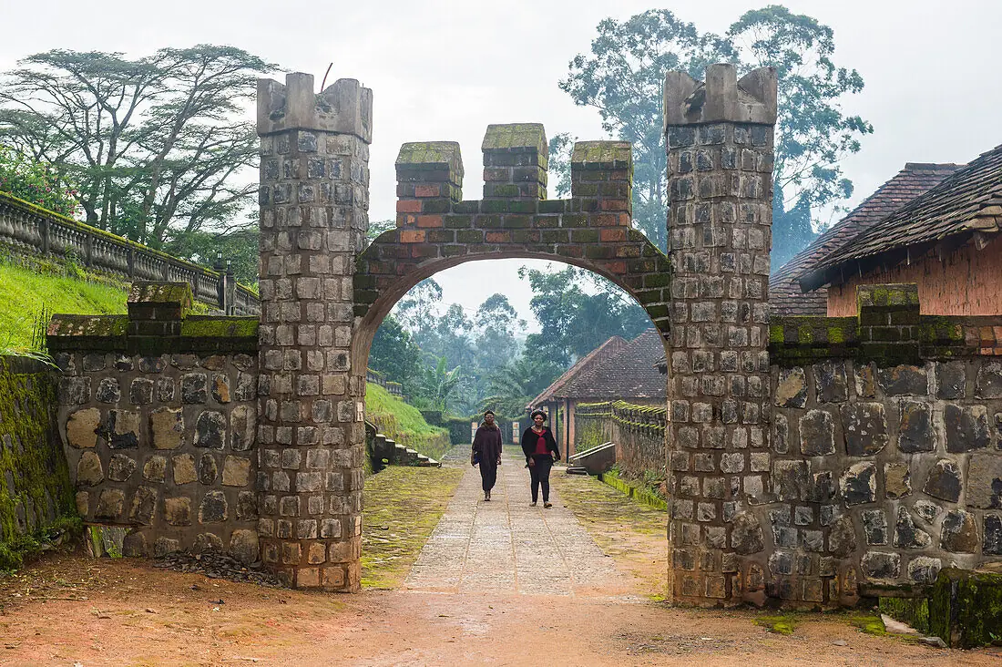 A stone archway with square towers opening onto a paved path, two people walking through it.