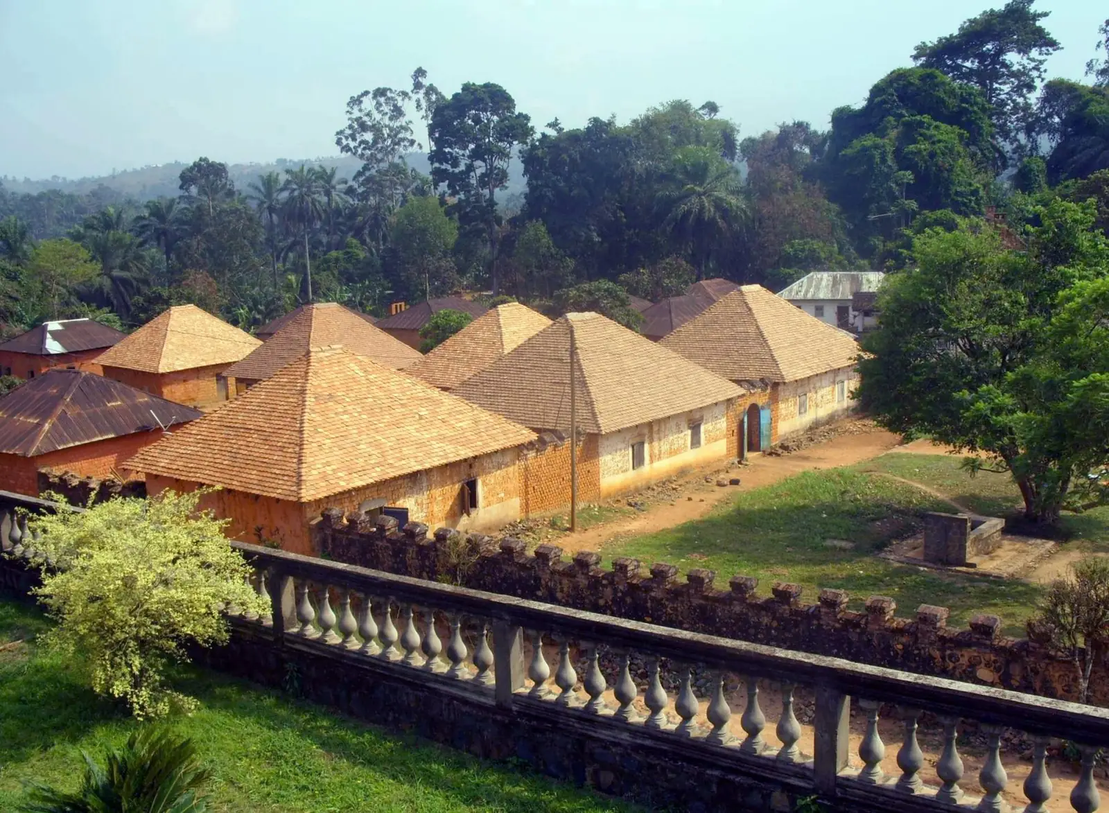 Rows of tile-roofed houses in the palace compound seen from a stone terrace, with palms and forested hills behind.