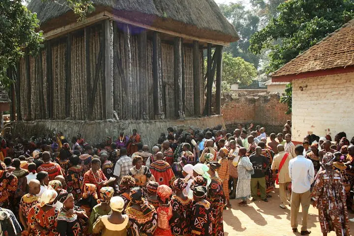A dense crowd in patterned cloth and caps gathered close against the wall of a thatched building.